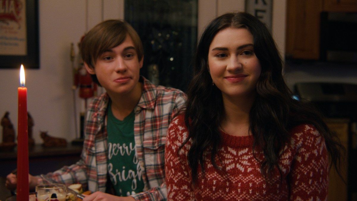 A teenage boy and a teenage girl wearing festive clothes sit next to each other at a dinner table.