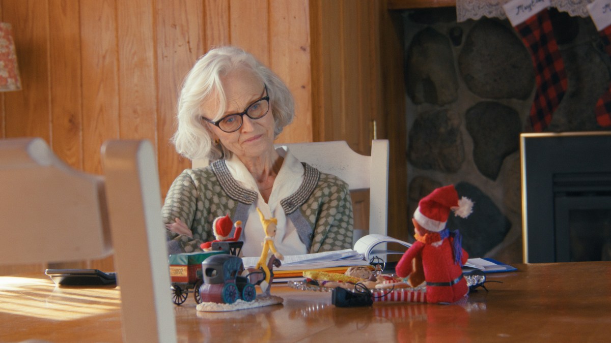 An old woman sits at a table and looks at a small stuffed elf.
