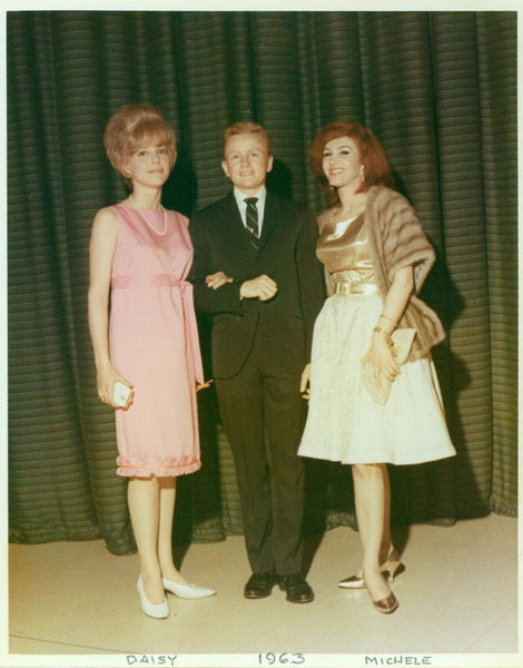 A short man in a black suit stands between two women in dresses. All three half various shades of red hair.