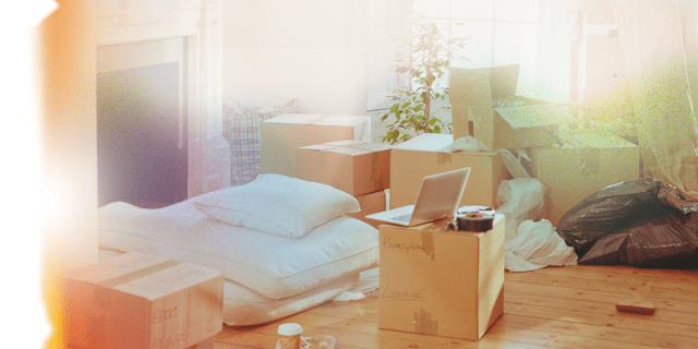 an image of a sleeping pad and pillow on the floor surrounded by boxes with a laptop balanced on the boxes. the photo has the effect of an overexposed piece of film on it so that the edges are blurred out and white with rainbows crossing across the image