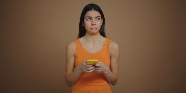 a woman in an orange tanktop holds a phone with a yellow case and grimaces as she seems to think about what to text. headline text