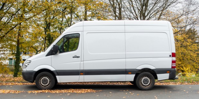 An empty white van in a parking lot with autumnal trees behind it.