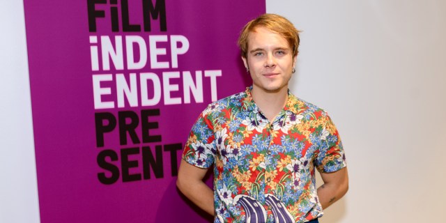 Vuk Lungulov-Klotz in a colorful shirt with a flower pattern stands in front of a purple sign that says Film Independent Presents