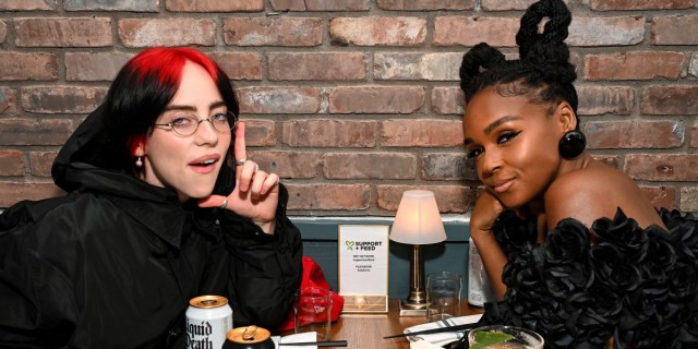 Billie Eilish sits across a table from Janelle Monáe. Billie rests her head in the shape of an L.