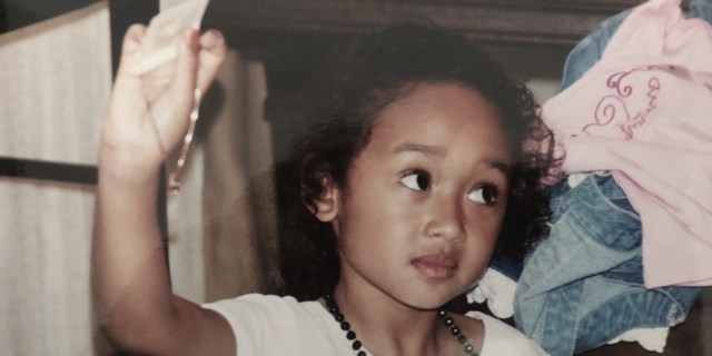A slightly desaturated childhood photo of a genderqueer child with curly hair wearing a white shirt and a beaded necklace glancing up with an uncertain face at a pink top.