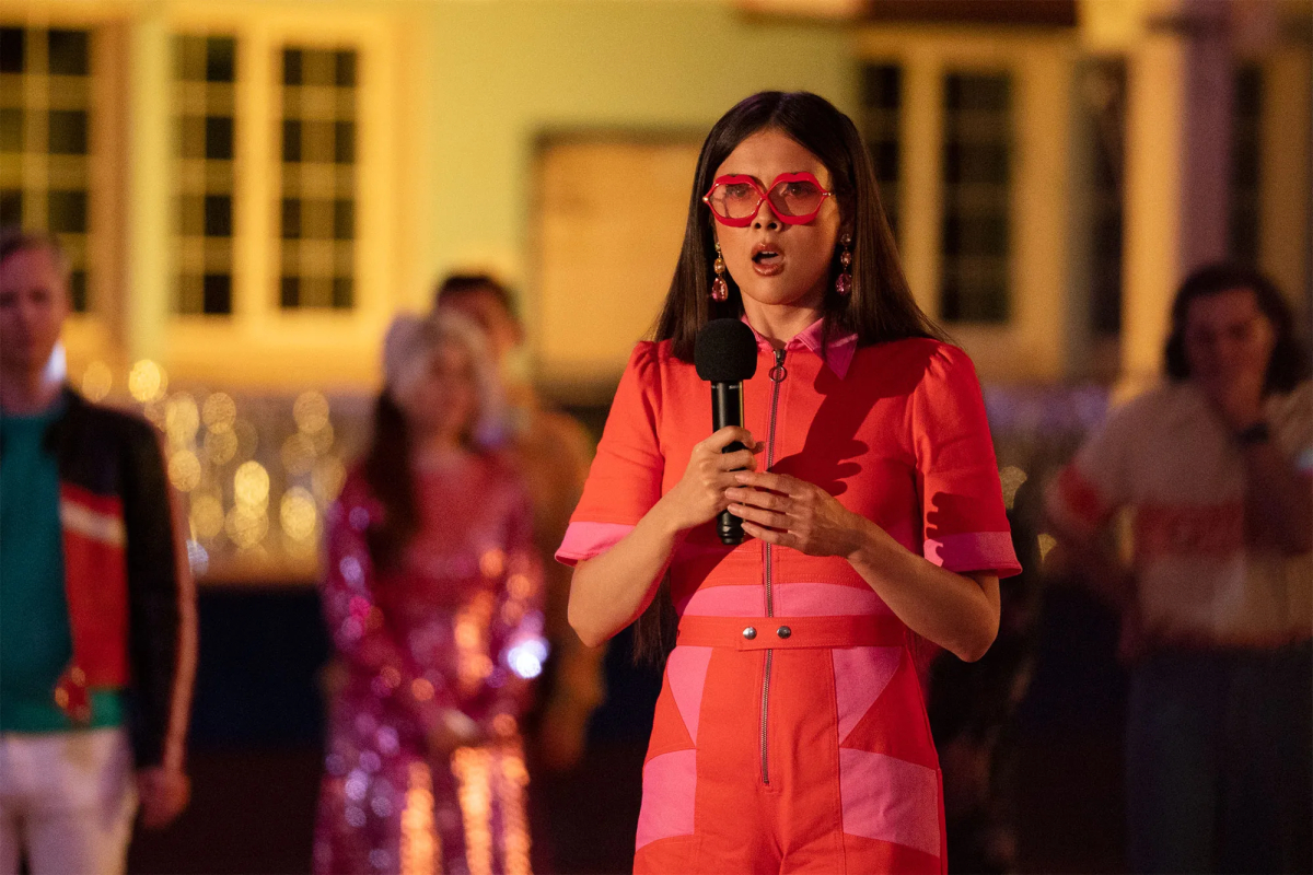 Patti Harrison in an orange and pink neon jumpsuit and big red lip glasses holds a mic and looks shocked