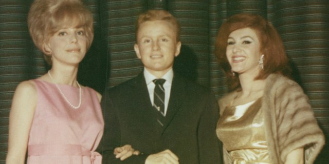A cropped close up of Reed Erickson, a short man in a black suit stands between two women in dresses. All three half various shades of red hair.