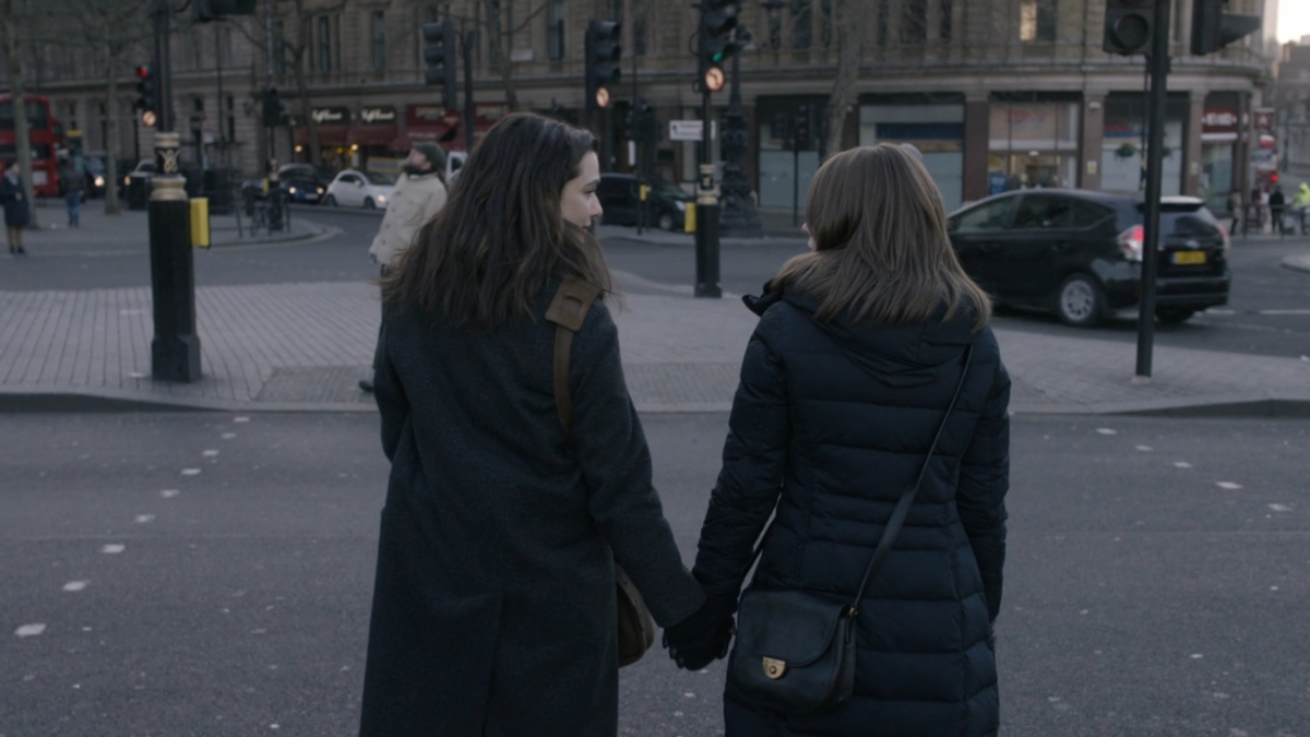 Ronit and Esti stand at a crosswalk in coats holding hands.