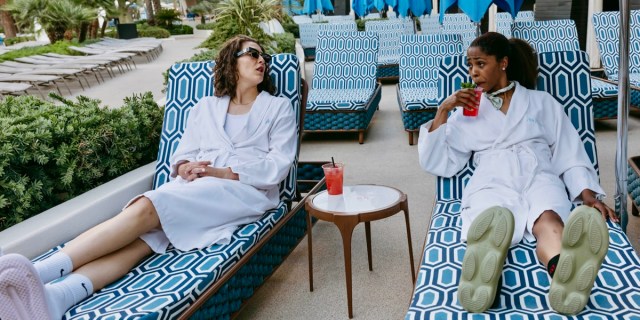 The Syd + TP Show: WNBA Stars Theresa Plaisance, a tall white brunette with curly hair, and Sydney Colson, a Black woman with hair in a ponytail, are both in bathrobes lounging on blue pool chairs.