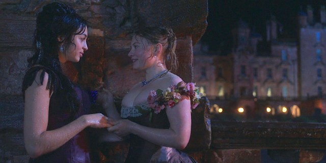 Josie Totah as Mabel and Mia Threapleton as Honoria hold hands against a wall drenched in rain water, a castle in the background.