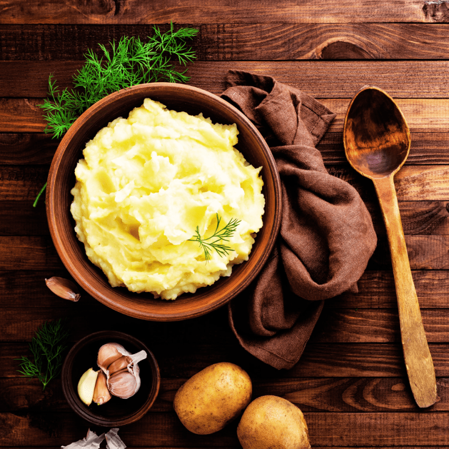 a photo of mashed potatoes in a bowl on a table with spoon, herbs and potatoes decorating the setup