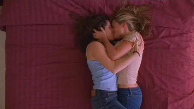 Two women hold each other while kissing on a red bed.