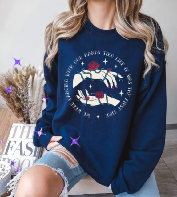 A blue sweatshirt with an illustration of two hands wrapped in roses with the circle text around it reading: we were dancing with our hands tied like it was the first time