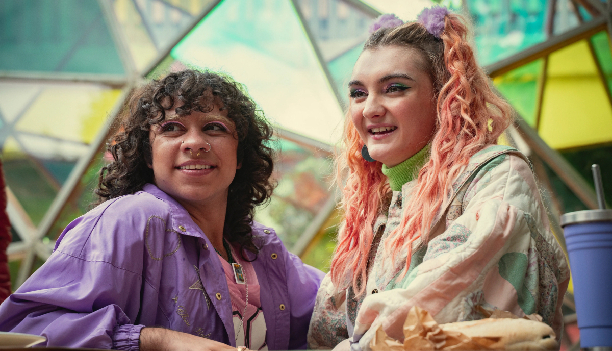 A transmasc in a purple jacket stands next to a trans girl in a light pastel jacket. They're both smiling and half colorful makeup.