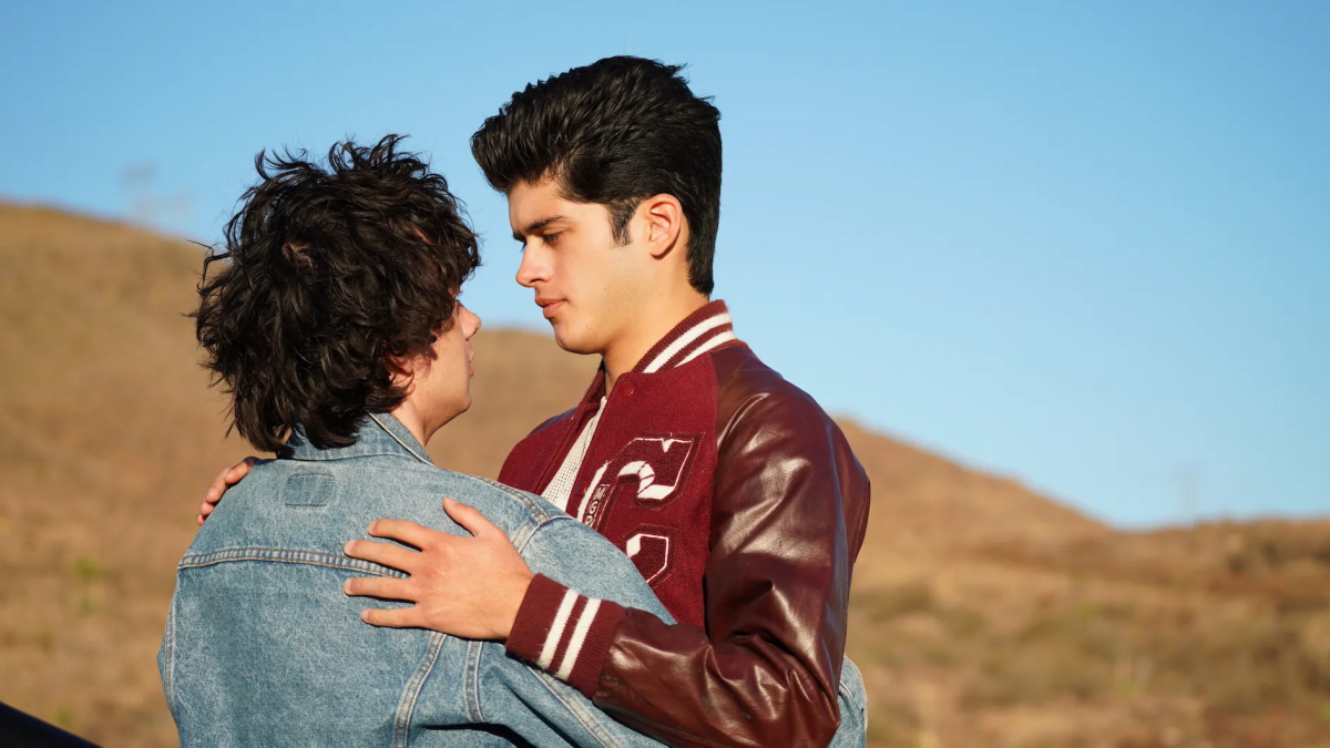 Two teen boys, one in a jean jacket, one in a letterman jacket, embrace with a desert terrain behind them.