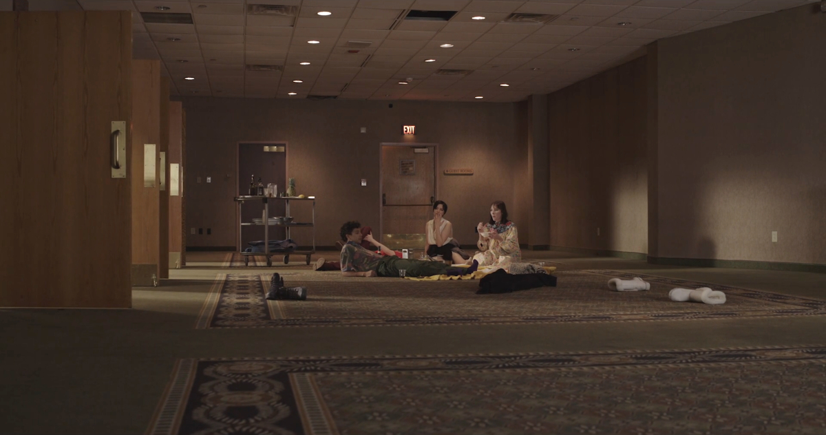 A wide shot of an empty hotel conference hall. Four queer people sit on the floor. 