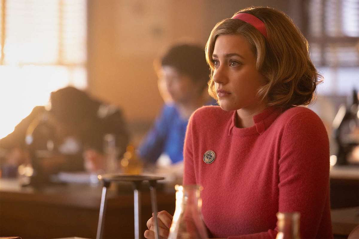 Lili Reinhart as Betty Cooper sits in class in a matching pink sweater and headband.