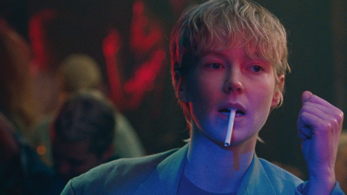 Close-up on a woman with short blonde hair at a bar, a cigarette dangles out of her mouth