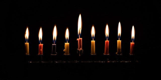 A menorah glows on the last night of Hanukkah, the candles almost burnt out, the background complete darkness.