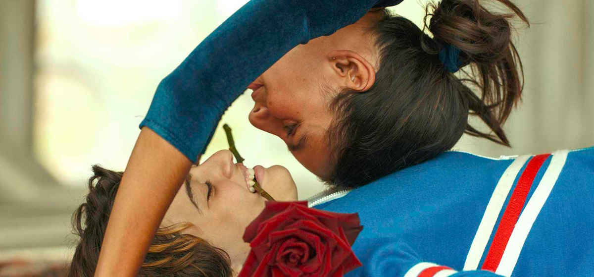 A transmasc child lies on the ground with a rose in their mouth as a girl does a headstand on their chest.