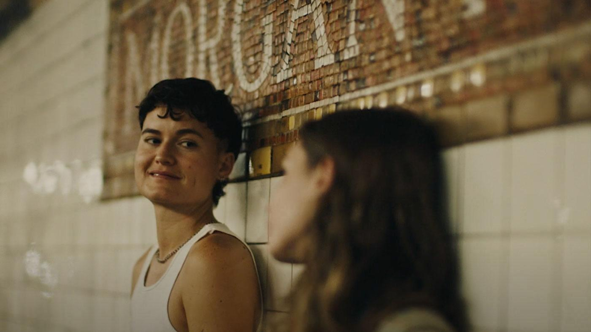 A transmasc in a white tank top glances down at his sister as they lean against the wall in the Morgan Ave subway stop.