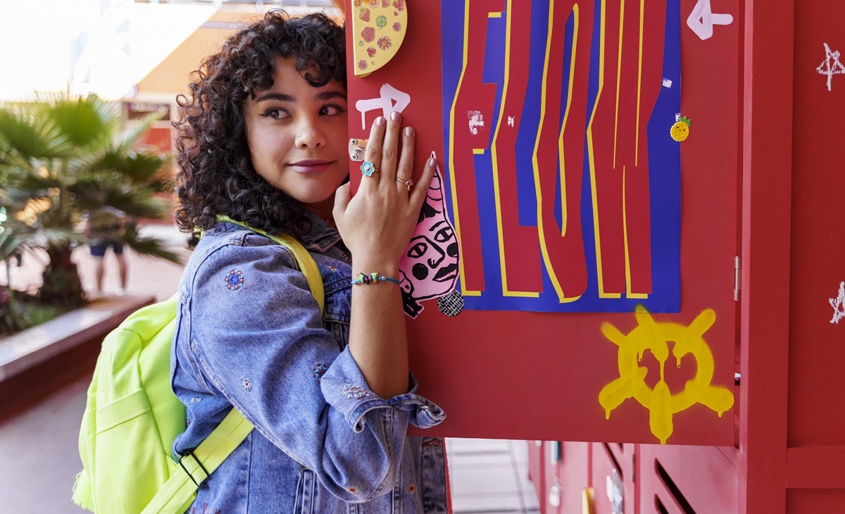 A teen girl wearing a jean jacket and a green neon backpack stands by her open red locker. 
