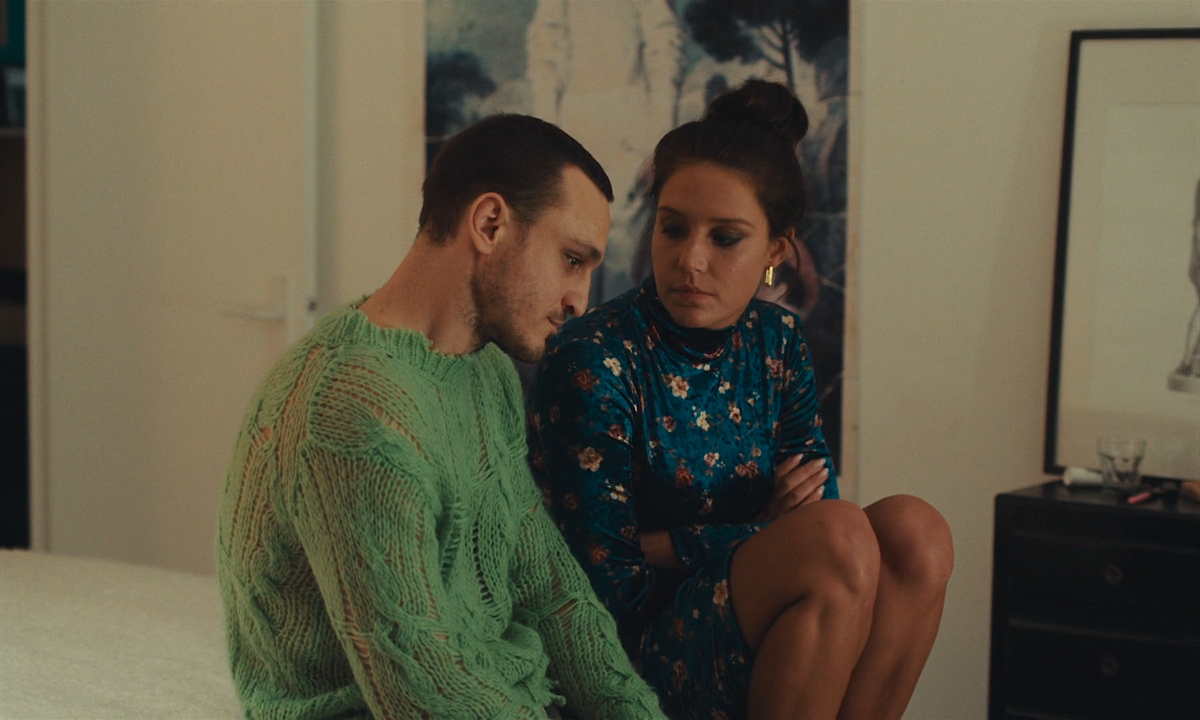 Best queer movies of 2023: Franz Rogowski and Adèle Exarchopoulos sit next to each other on a bed, Rogowski in a green sweater, Exarchopoulos in a blue dress.