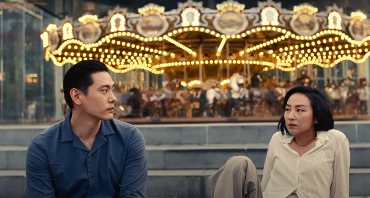 Greta Lee and Teo Yoo sit on stairs in front of a carousel and look at one another. 