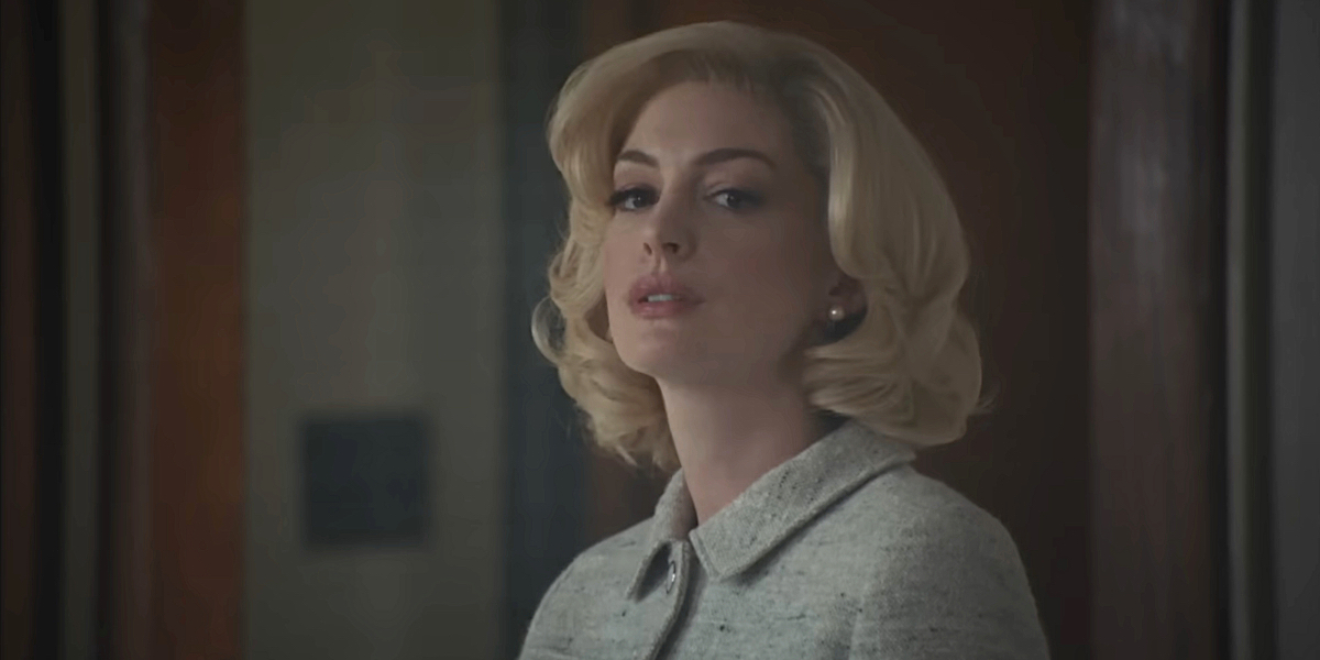 Eileen: Anne Hathaway in a blonde wig purses her lips and looks toward the camera.