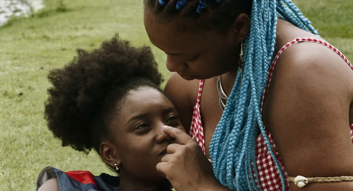 Best queer movies of 2023: A young woman leans in the lap of another young woman. The second woman puts a finger on the first's nose as her blue braids cascade down. 
