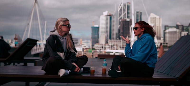 Kristie Mewis and Lynn Williams talk, while sitting on a table with the New Zealand city skyline behind them.