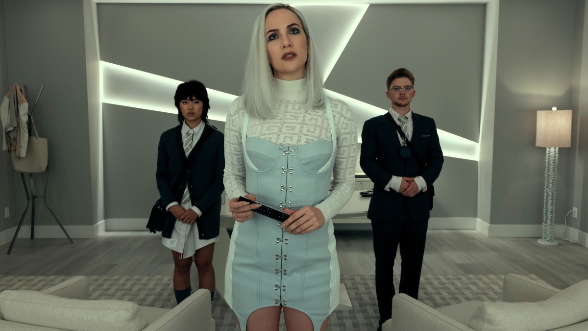 Kate Siegel in a white wig, white turtleneck sweater, and a light blue denim jumper stands between her two assistants in suits.