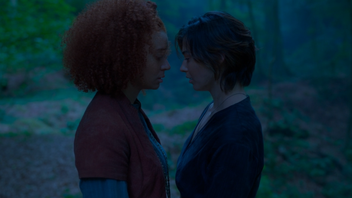 Two girls, one with short hair, one with longer curly hair, stand facing each other in the woods.