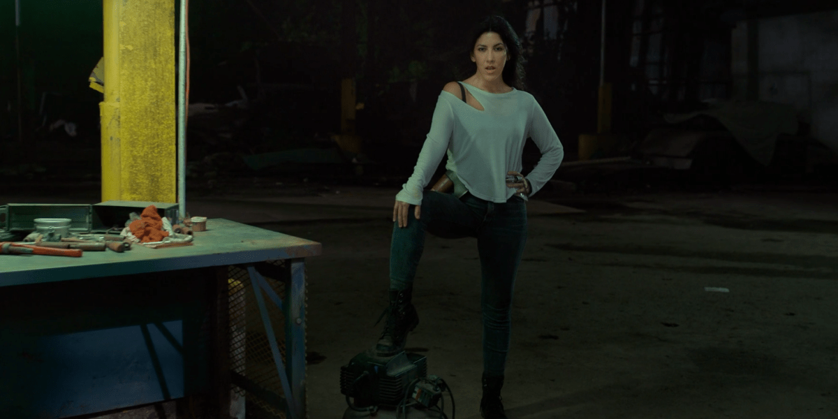 Stephanie Beatrix in a loose blue top and tight black pants lifts one leg up onto a car engine. 