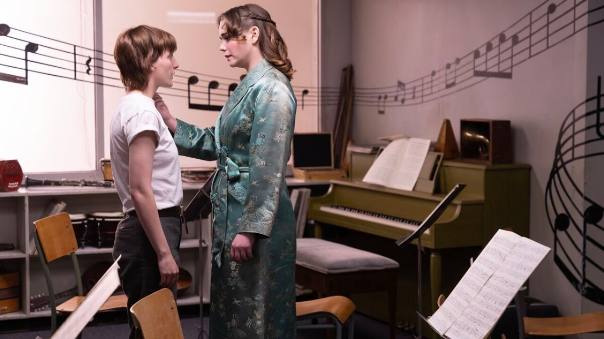 A butch in a white shirt and femme in a teal robe look at each other in a school music room.