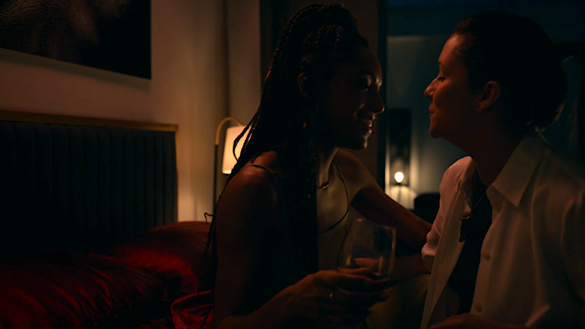 Two women shrouded in darkness smile at each other while sitting on a bed.