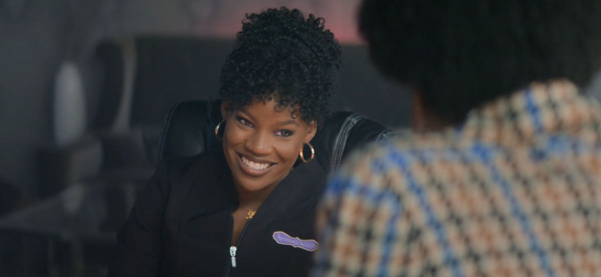 Brittany Adebumola as Malaika smiles wearing a black jumpsuit and gold earrings.