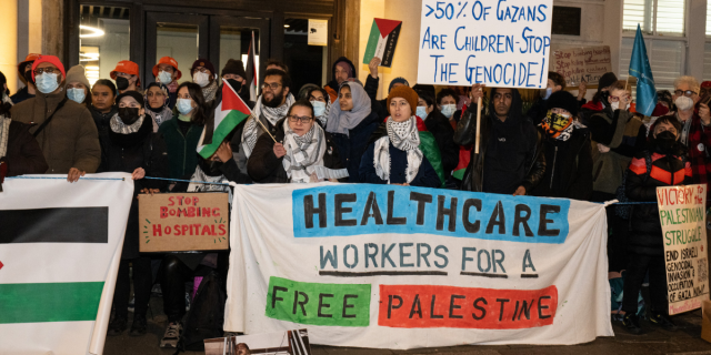 protesters gather to protest Israel's genocide of Palestine, and a group holds a banner that says HEALTHCARE WORKERS FOR A FREE PALESTINE