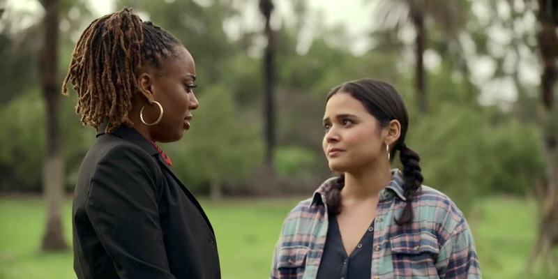 Tracy confronts Malika for half-assing her participation in the retreat activities. Malika is on the left, wearing a black blazer over her flowy red dress. Tracy is on the right, wearing a flannel shirt over a v-neck top.
