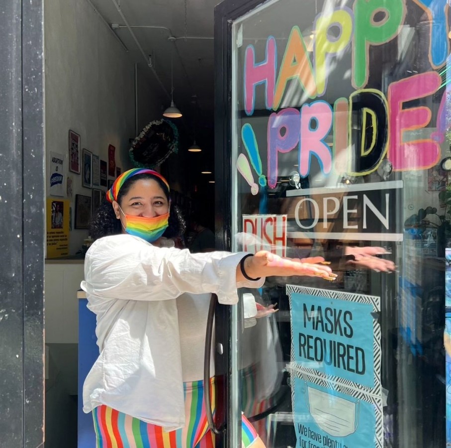 a worker owner at Bluestockings in a mask welcomes folks into the store which has a HAPPY PRIDE sign outside it