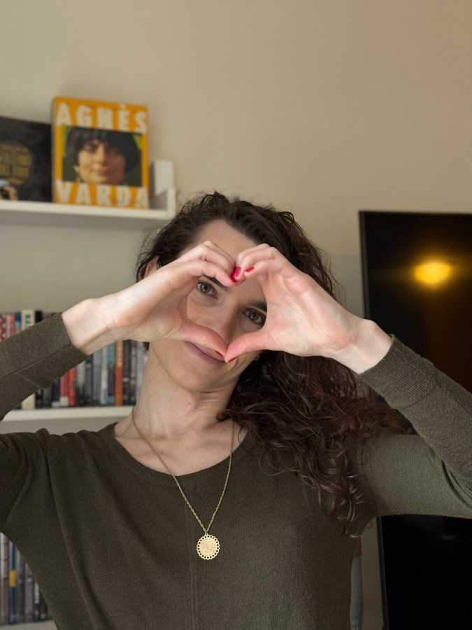 Drew, a white woman with long curly hair, holds her hands up in a heart shape