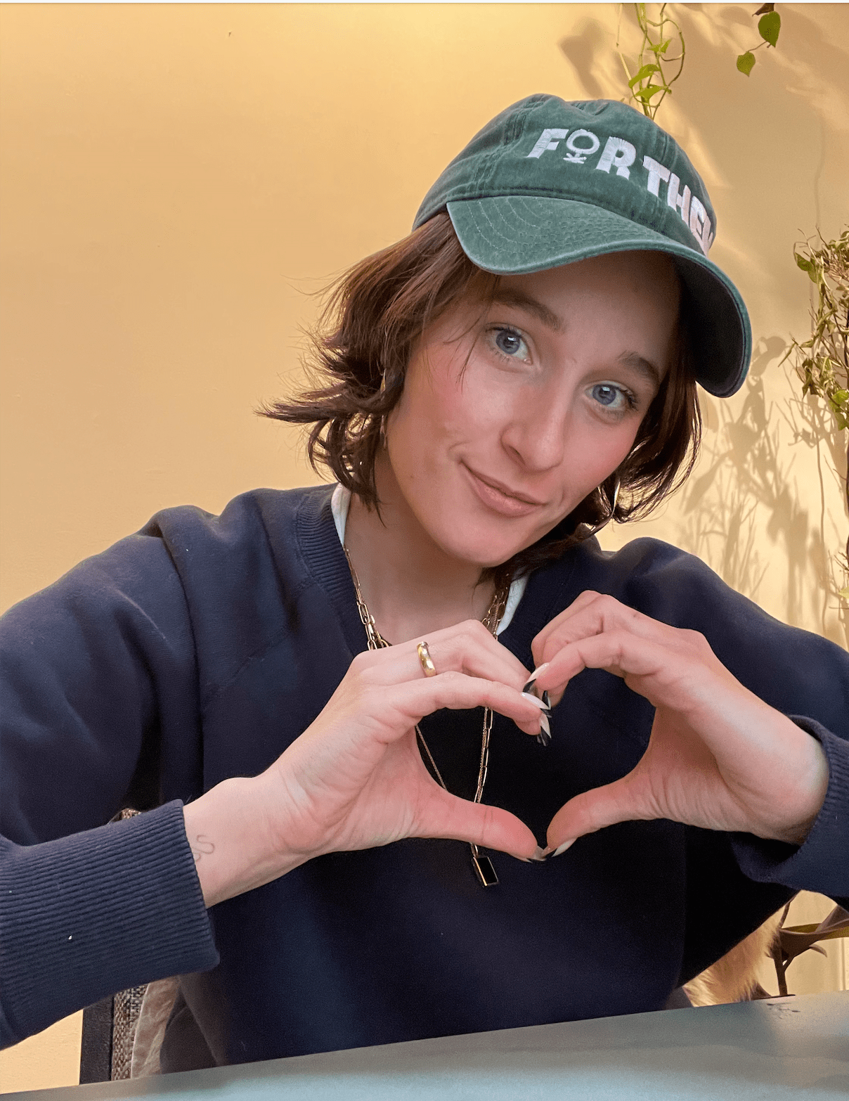 Em, a white woman with short wavy brown hair wearing a for them baseball cap holds her hands up in a heart shape
