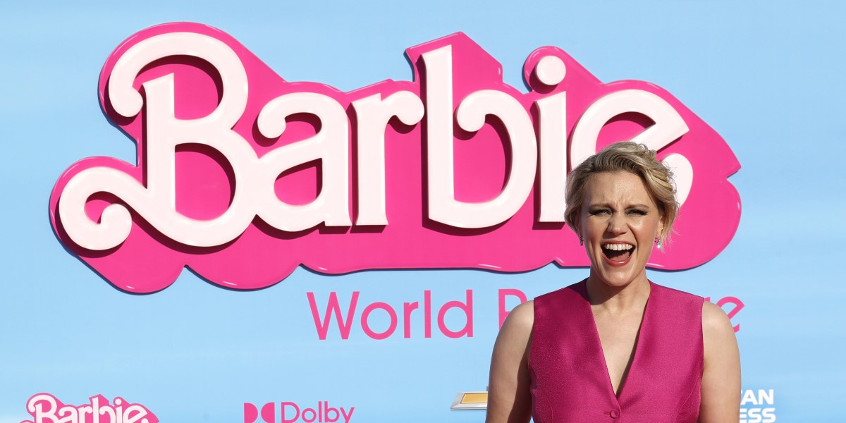 Kate McKinnon dressed in pink in front of the Barbie logo