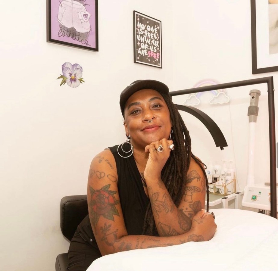 A queer Black woman with tattoos on her arms rests her head on her hand as she leans against a facial table.