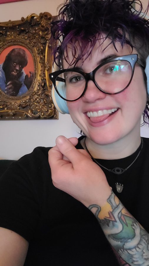 nico, a white genderfluid human with short purple curly hair and glasses holds their fingers up in a heart shape