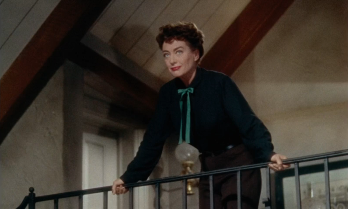 Joan Crawford in a button down and pants leans over a railing.