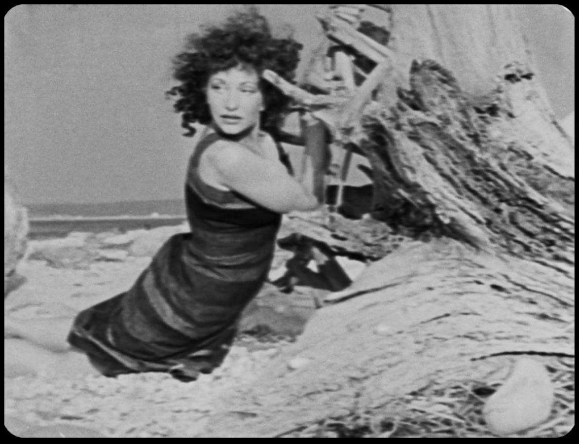 Maya Deren grips a tree on a beach