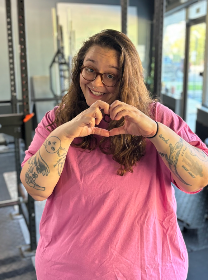 stef, a white nonbinary human with glasses and long brown hair, holds their hands up in a heart shape while at the gym