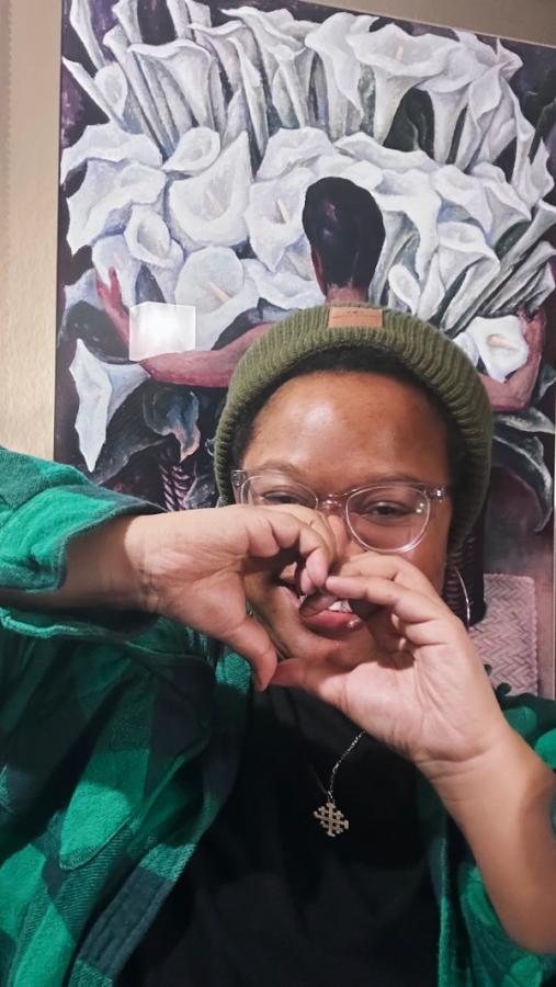 carmen, a Black woman wearing a beanie and clear glasses, holds up her hands in a heart shape