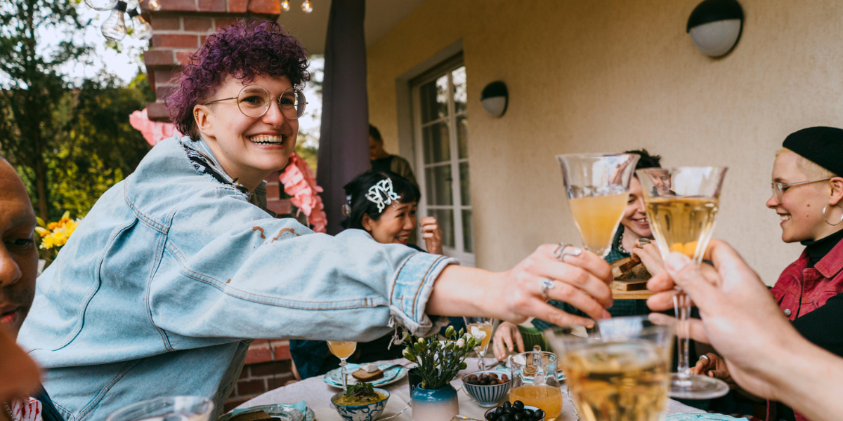 a queer person with purple curly hair toasts with a glass of champagne and wears a denim jacket